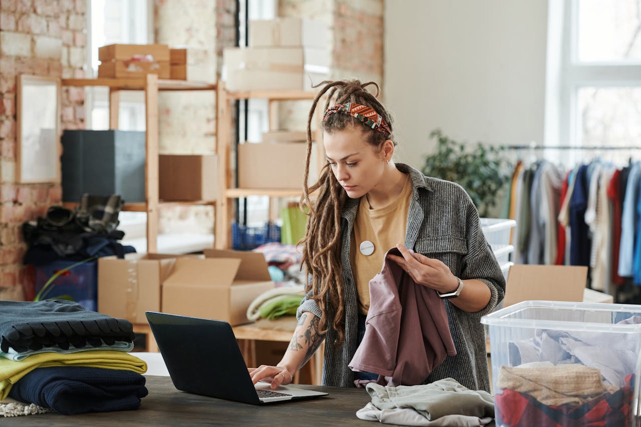 Woman with dreadlocks using a laptop while sorting clothes in a loft for volunteer work.
